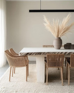 A modern dining room featuring a bleached oak dining table with a live edge, woven rattan dining chairs, and a large, textured cream area rug. A matte black linear pendant light hangs above, and a minimalist ceramic vase with dried pampas grass adorns the table.