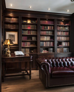 A traditional library featuring dark walnut built-in bookshelves filled with books, a deep oxblood leather Chesterfield sofa, and a classic banker's lamp on a mahogany desk. Warm ambient lighting from recessed ceiling lights and a bronze chandelier creates a refined glow. — close-up of textures and materials