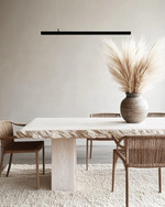 A modern dining room featuring a bleached oak dining table with a live edge, woven rattan dining chairs, and a large, textured cream area rug. A matte black linear pendant light hangs above, and a minimalist ceramic vase with dried pampas grass adorns the table. — alternate angle