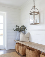 A bright coastal mudroom featuring a white shiplap wall, a light-toned reclaimed wood bench, oversized woven rattan baskets for storage, and a striking glass-encased brushed nickel lantern pendant light illuminating the space. — alternate angle