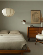 A Japandi bedroom blending vintage and modern, featuring a low oak bed, a mid-century modern teak dresser, a modern rice paper pendant light, and a vintage spindle-back chair with a sheepskin throw, all in muted tones of soft grey, beige, and sage green. — alternate angle