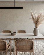 A modern dining room featuring a bleached oak dining table with a live edge, woven rattan dining chairs, and a large, textured cream area rug. A matte black linear pendant light hangs above, and a minimalist ceramic vase with dried pampas grass adorns the table. — styling and décor detail