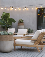 A minimalist boho deck featuring light teak wood lounge chairs with off-white cushions, subtle woven rattan accents, and large concrete planters holding a Fiddle-Leaf Fig and various succulents, all under warm string lights. — alternate angle