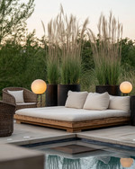 A Scandinavian poolside area featuring a large reclaimed teak daybed with cream linen cushions, oversized woven rattan armchairs, and matte black metal planters with tall Horsetail Reed plants, all under the soft glow of spherical outdoor lanterns. — close-up of textures and materials