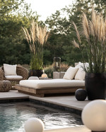 A Scandinavian poolside area featuring a large reclaimed teak daybed with cream linen cushions, oversized woven rattan armchairs, and matte black metal planters with tall Horsetail Reed plants, all under the soft glow of spherical outdoor lanterns. — styling and décor detail
