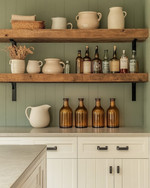 Calm farmhouse home bar featuring warm white shaker cabinets, a honed light grey soapstone countertop, and open reclaimed wood shelving. Amber glass bottles, ceramic pitchers, and matte black hardware complement the serene sage green accent wall.