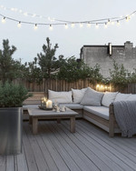 A serene Scandinavian deck with light-toned wooden flooring, a low-profile wooden lounge set with off-white cushions and a chunky knit grey wool throw, minimalist galvanized steel planters with evergreens, and soft string lights overhead. — styling and décor detail