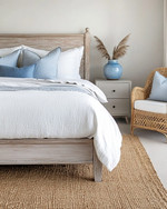 Coastal bedroom with a calming color palette featuring a whitewashed wooden bed frame, crisp white linen bedding, and soft blue throw pillows. A natural jute rug grounds the space, while a rattan armchair and a light grey nightstand with a ceramic vase complete the serene atmosphere.