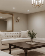 A refined traditional basement living area featuring light greige walls, a tailored neutral linen Chesterfield sofa, and a simple dark wood coffee table. Recessed lighting and a classic brass-finished chandelier illuminate the space, complemented by a large unframed mirror and minimalist decor.
