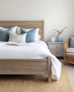 Coastal bedroom with a calming color palette featuring a whitewashed wooden bed frame, crisp white linen bedding, and soft blue throw pillows. A natural jute rug grounds the space, while a rattan armchair and a light grey nightstand with a ceramic vase complete the serene atmosphere. — close-up of textures and materials