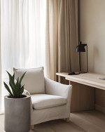 A serene Japandi home office with a pale oak desk, a minimalist matte black desk lamp, and an off-white linen armchair. Natural light streams through sheer curtains, illuminating a small snake plant in a raw concrete planter. — alternate angle
