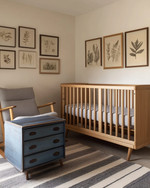 A modern baby and kids room featuring a natural maple crib, a mid-century modern rocking chair with grey linen upholstery, and a vintage wooden toy chest in faded blue. The walls are painted in a warm off-white, complemented by a subtle striped rug and a collection of vintage framed botanical prints. — close-up of textures and materials