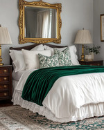 A traditional bedroom featuring a bed dressed in white and cream sateen bedding, layered with a deep emerald green velvet throw and patterned decorative pillows. A large ornate gold-framed mirror hangs above a dark wood dresser, flanked by classic brass table lamps with pleated fabric shades. A muted Persian-style rug covers the floor. — alternate angle