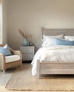 Coastal bedroom with a calming color palette featuring a whitewashed wooden bed frame, crisp white linen bedding, and soft blue throw pillows. A natural jute rug grounds the space, while a rattan armchair and a light grey nightstand with a ceramic vase complete the serene atmosphere. — alternate angle