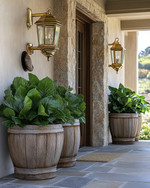 A coastal driveway featuring large weathered teak planters filled with lush green foliage, flanked by elegant nautical brass lanterns mounted on stone pillars, leading to a front door with a pewter shell door knocker. — close-up of textures and materials