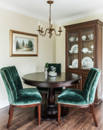 A small traditional dining room featuring a round dark wood pedestal table, upholstered dining chairs in a deep emerald green velvet, and a bronze chandelier hanging above. The walls are painted in a warm cream, and a narrow, glass-front china cabinet stands against one wall, displaying classic dinnerware. — styling and décor detail