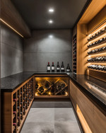 Modern wine cellar with warm grey walls, rift-sawn oak wine racks, and a honed black granite tasting counter. Integrated LED strip lighting illuminates the wine bottles, while recessed spotlights provide ambient glow.