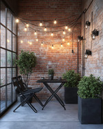 A compact industrial patio featuring a foldable black metal bistro set, string lights with exposed Edison bulbs, and potted green plants in dark grey concrete planters, set against an exposed brick wall.