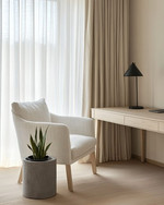 A serene Japandi home office with a pale oak desk, a minimalist matte black desk lamp, and an off-white linen armchair. Natural light streams through sheer curtains, illuminating a small snake plant in a raw concrete planter. — close-up of textures and materials
