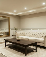 A refined traditional basement living area featuring light greige walls, a tailored neutral linen Chesterfield sofa, and a simple dark wood coffee table. Recessed lighting and a classic brass-finished chandelier illuminate the space, complemented by a large unframed mirror and minimalist decor. — alternate angle