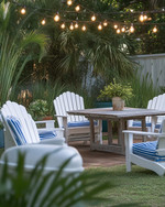 A serene coastal backyard featuring white Adirondack chairs, a weathered teak dining table, and blue and white striped outdoor cushions under strung Edison bulb lights, surrounded by lush green ornamental grasses and potted sago palms.