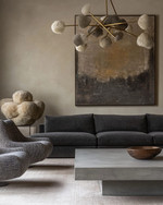 Modern great room featuring a low-profile charcoal grey sectional sofa, a large polished concrete coffee table, and a pair of bouclé accent chairs. A dramatic brushed brass chandelier hangs above, complemented by a large abstract painting on a light greige wall. — alternate angle