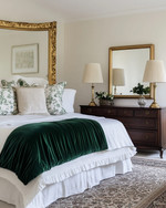 A traditional bedroom featuring a bed dressed in white and cream sateen bedding, layered with a deep emerald green velvet throw and patterned decorative pillows. A large ornate gold-framed mirror hangs above a dark wood dresser, flanked by classic brass table lamps with pleated fabric shades. A muted Persian-style rug covers the floor. — styling and décor detail