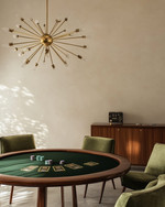 A mid-century modern game room featuring a walnut poker table with a dark green felt top, surrounded by olive velvet low-slung armchairs, illuminated by a brass Sputnik chandelier, with a teak credenza against a soft ivory wall. — alternate angle