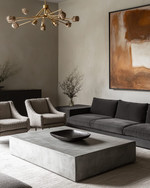 Modern great room featuring a low-profile charcoal grey sectional sofa, a large polished concrete coffee table, and a pair of bouclé accent chairs. A dramatic brushed brass chandelier hangs above, complemented by a large abstract painting on a light greige wall.