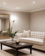 A refined traditional basement living area featuring light greige walls, a tailored neutral linen Chesterfield sofa, and a simple dark wood coffee table. Recessed lighting and a classic brass-finished chandelier illuminate the space, complemented by a large unframed mirror and minimalist decor. — styling and décor detail