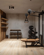 A clean rustic home gym featuring light reclaimed wood flooring, a smooth concrete accent wall, a simple black wrought iron shelf, and minimalist workout equipment with wooden and leather details, bathed in soft ambient lighting. — alternate angle