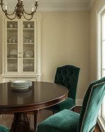 A small traditional dining room featuring a round dark wood pedestal table, upholstered dining chairs in a deep emerald green velvet, and a bronze chandelier hanging above. The walls are painted in a warm cream, and a narrow, glass-front china cabinet stands against one wall, displaying classic dinnerware. — close-up of textures and materials