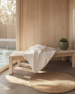 A compact Scandinavian poolhouse interior featuring light birch wood furniture, a white linen throw on a simple bench, and a small round jute rug. A ceramic planter with a green succulent sits on a minimalist shelf, reflecting natural light from a large window.