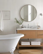 A vintage modern coastal bathroom featuring a white clawfoot tub, a light oak floating vanity, and a round frameless mirror, accented with polished nickel fixtures and a jute bath mat. — alternate angle
