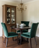 A small traditional dining room featuring a round dark wood pedestal table, upholstered dining chairs in a deep emerald green velvet, and a bronze chandelier hanging above. The walls are painted in a warm cream, and a narrow, glass-front china cabinet stands against one wall, displaying classic dinnerware.