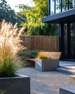 A Scandinavian front yard featuring a pea gravel path, large concrete planters with ornamental grasses, a low woven willow fence, and a slatted wooden bench by the matte black front door.