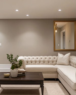 A refined traditional basement living area featuring light greige walls, a tailored neutral linen Chesterfield sofa, and a simple dark wood coffee table. Recessed lighting and a classic brass-finished chandelier illuminate the space, complemented by a large unframed mirror and minimalist decor. — close-up of textures and materials
