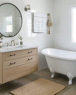 A vintage modern coastal bathroom featuring a white clawfoot tub, a light oak floating vanity, and a round frameless mirror, accented with polished nickel fixtures and a jute bath mat. — styling and décor detail