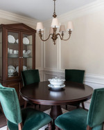 A small traditional dining room featuring a round dark wood pedestal table, upholstered dining chairs in a deep emerald green velvet, and a bronze chandelier hanging above. The walls are painted in a warm cream, and a narrow, glass-front china cabinet stands against one wall, displaying classic dinnerware. — alternate angle