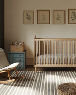 A modern baby and kids room featuring a natural maple crib, a mid-century modern rocking chair with grey linen upholstery, and a vintage wooden toy chest in faded blue. The walls are painted in a warm off-white, complemented by a subtle striped rug and a collection of vintage framed botanical prints. — alternate angle