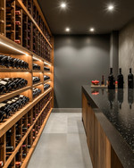 Modern wine cellar with warm grey walls, rift-sawn oak wine racks, and a honed black granite tasting counter. Integrated LED strip lighting illuminates the wine bottles, while recessed spotlights provide ambient glow. — alternate angle