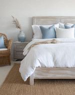 Coastal bedroom with a calming color palette featuring a whitewashed wooden bed frame, crisp white linen bedding, and soft blue throw pillows. A natural jute rug grounds the space, while a rattan armchair and a light grey nightstand with a ceramic vase complete the serene atmosphere. — styling and décor detail