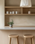 A Japandi home bar featuring light oak cabinetry, a honed light grey quartz countertop, and minimalist open shelving displaying simple ceramic mugs and a small green plant. Two backless wooden stools are neatly tucked beneath the counter, and soft, diffused light emanates from a simple linen pendant.