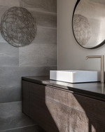 A modern powder room features a floating dark wood vanity with an integrated white quartz sink and brushed nickel faucet, set against large format charcoal grey porcelain tiles. A simple round mirror and abstract metal wall art complete the sleek, resilient design. — styling and décor detail