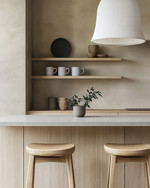 A Japandi home bar featuring light oak cabinetry, a honed light grey quartz countertop, and minimalist open shelving displaying simple ceramic mugs and a small green plant. Two backless wooden stools are neatly tucked beneath the counter, and soft, diffused light emanates from a simple linen pendant. — alternate angle
