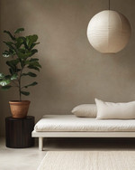 A serene Japandi poolhouse interior featuring a low-slung white oak daybed with natural linen cushions, a dark wenge wood side table, and a large, round paper lantern pendant light. The walls are a soft greige, complemented by a subtle textured rug in cream and a tall, slender fiddle leaf fig plant in a terracotta pot. — alternate angle