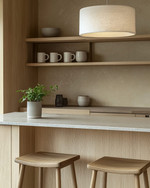 A Japandi home bar featuring light oak cabinetry, a honed light grey quartz countertop, and minimalist open shelving displaying simple ceramic mugs and a small green plant. Two backless wooden stools are neatly tucked beneath the counter, and soft, diffused light emanates from a simple linen pendant. — close-up of textures and materials