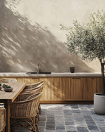 Japandi outdoor kitchen featuring untreated teak cabinetry, honed concrete countertops, and bluestone pavers. Minimalist rattan dining chairs surround a solid wood table, with an olive tree in an unglazed ceramic planter providing a natural accent. — alternate angle