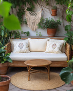 A cozy boho patio featuring a low-profile rattan loveseat with cream cushions, layered with macrame and embroidered pillows. A round jute rug sits beneath a small wooden coffee table, surrounded by various potted plants in terracotta and ceramic planters, including a trailing Pothos in a macrame hanger. — close-up of textures and materials