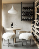 A small Scandinavian wine cellar with light ash wood shelving, a light gray concrete floor, and matte black metal wine racks. A minimalist white pendant light hangs over a small, round light wood tasting table with two simple stools covered in white sheepskin throws. — alternate angle